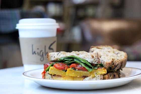 A veggie sandwich on a plate with a high5 coffee cup behind