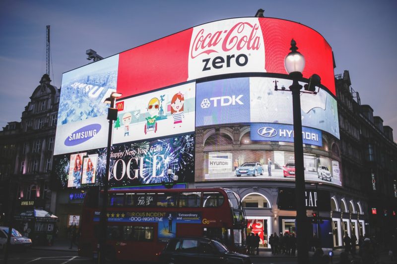 bright screens at night with people and a red double decker bus 